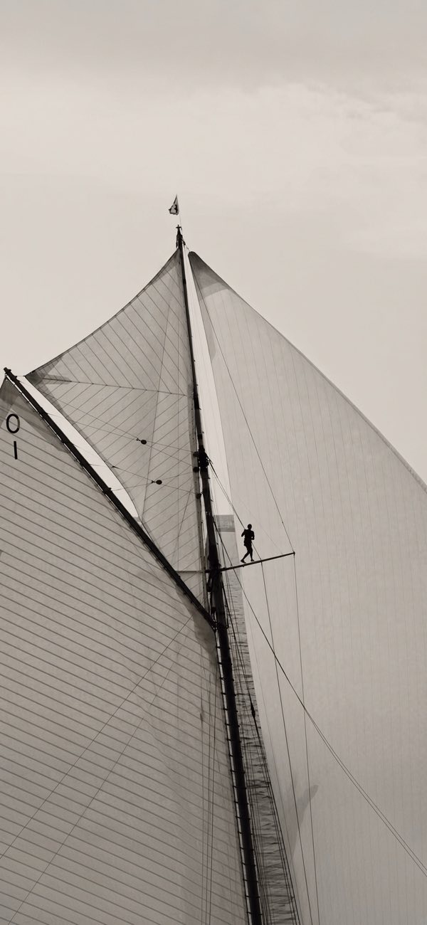 Looking up a gaff rig: sails, mast, a sailor on the spreader, in sand tones